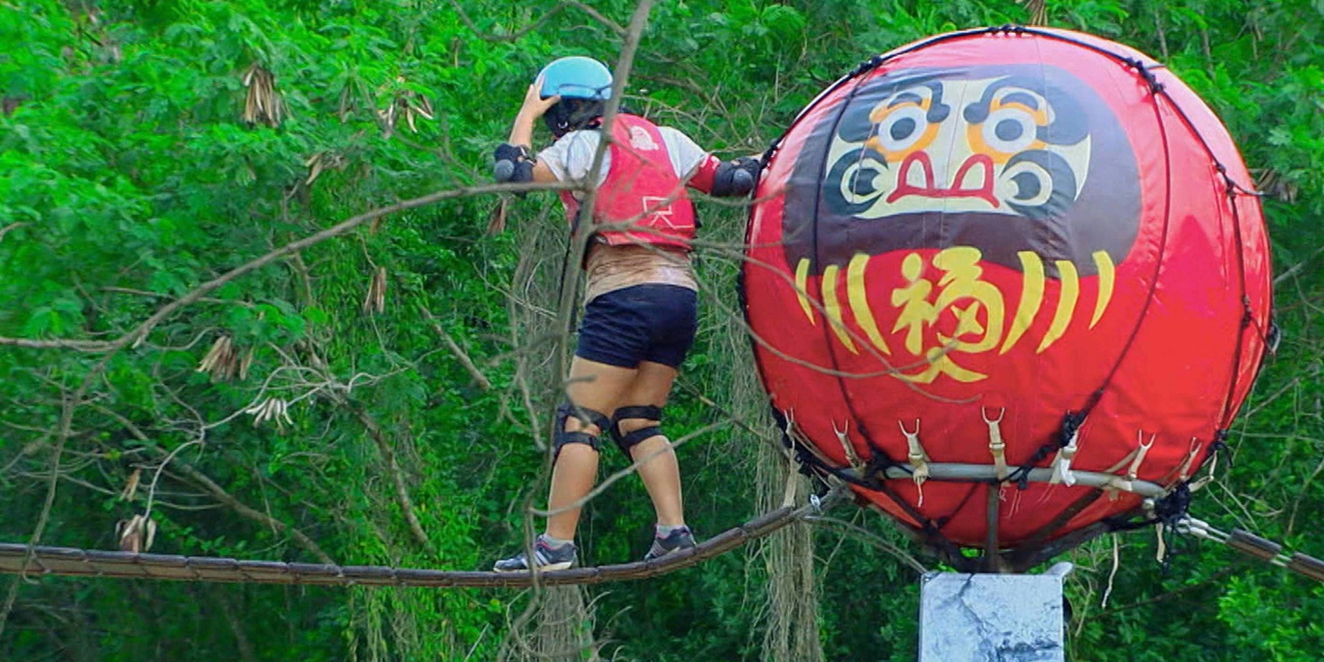 A contestant begins to cross a narrow bridge after climbing over a giant daruma